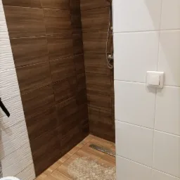 Kabina prysznicowa z brązowymi płytkami imitującymi drewno na ścianach i podłodze, białe płytki na sąsiedniej ścianie, odpływ liniowy w podłodze i widoczna słuchawka prysznicowa.