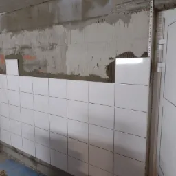 Białe, kwadratowe płytki częściowo ułożone na ścianie z surowym tynkiem, widoczne niedociągnięcia i resztki zaprawy, plastikowy narożnik ochronny przy futrynie drzwi.