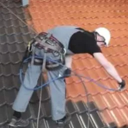 Pracownik w uprzęży, z kaskiem na głowie, częściowo pomalowany dach z dachówki ceramicznej, po lewej stronie kolor czarny, po prawej czerwony.