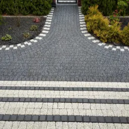 Ułożona z kostki brukowej ścieżka w ogrodzie, z dekoracyjnym obramowaniem z białych elementów, prowadząca do bramy z zielonymi krzewami po bokach.