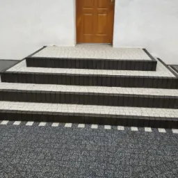 Zewnętrzne schody wejściowe z ciemnymi obrzeżami i jasnymi płytkami prowadzące do drewnianych drzwi wejściowych.