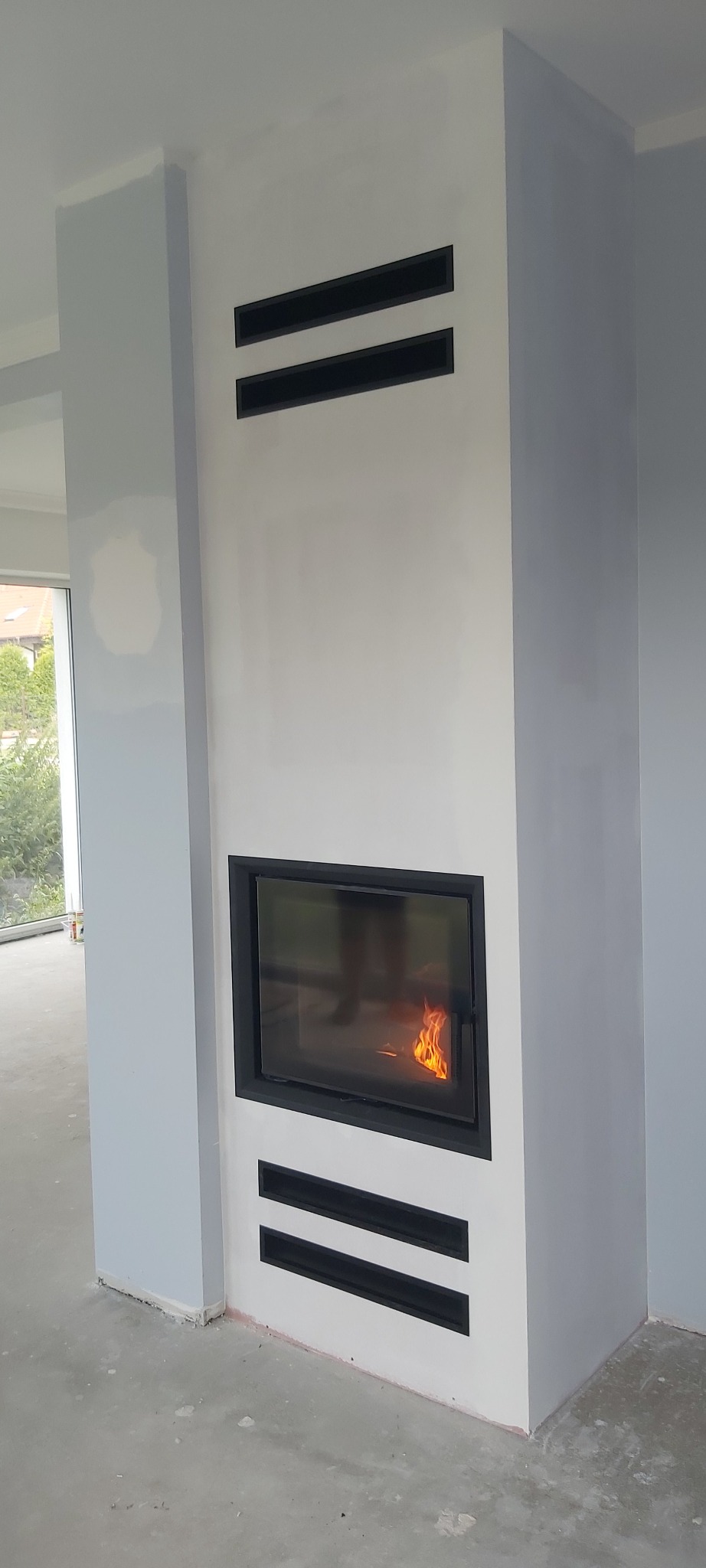 Nowoczesny wkład kominkowy z płomieniami, obudowany białą konstrukcją z czarnymi, poziomymi szczelinami wentylacyjnymi, widoczny w surowym stanie wykończeniowym wnętrza.