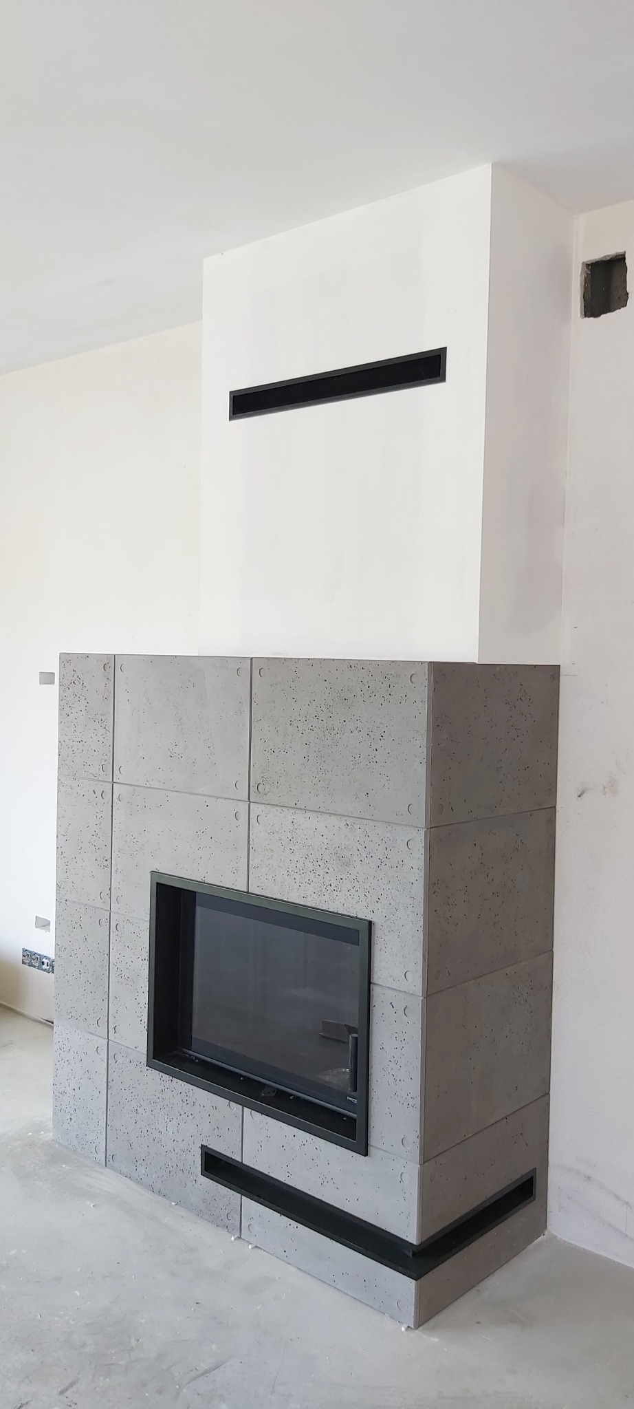 Nowoczesny kominek obłożony płytami z betonu architektonicznego z czarną ramą paleniska i poziomymi szczelinami wentylacyjnymi, w surowym, minimalistycznym wnętrzu.