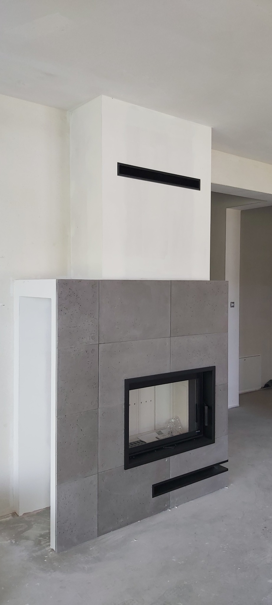 Nowoczesna obudowa wkładu kominkowego z szarego betonu architektonicznego, z czarnym obramowaniem paleniska i poziomym wlotem powietrza nad nim, w surowym wnętrzu.