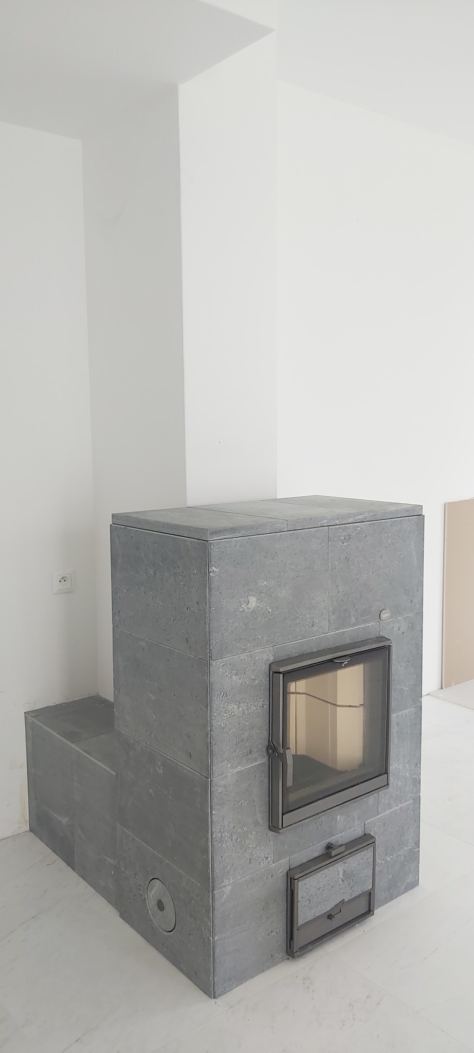 Nowoczesny piec kaflowy w odcieniach szarości, z żeliwnymi drzwiczkami i małym otworem na popiół, w surowym wnętrzu z białymi ścianami i marmurową podłogą.