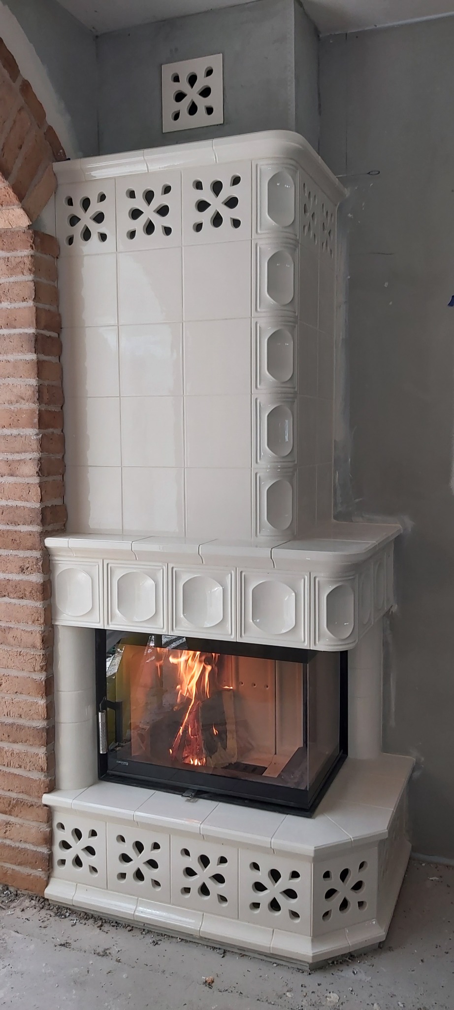 Biały, kaflowy piec z paleniskiem, płomienie widoczne przez szybę, obok fragment ceglanej ściany, surowy stan wykończenia wnętrza.