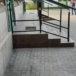 Denys Boiko - Zewnętrzne schody wejściowe do budynku z brązowymi płytkami, czarną metalową poręczą i szarą kostką brukową na chodniku.