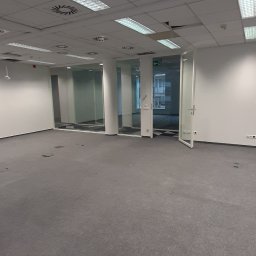Denys Boiko - Pusta przestrzeń biurowa z szarą wykładziną, białymi ścianami, podwieszanym sufitem z lampami i systemem wentylacji, oraz szklanymi ścianami działowymi oddzielającymi kolejne pomieszczenia.