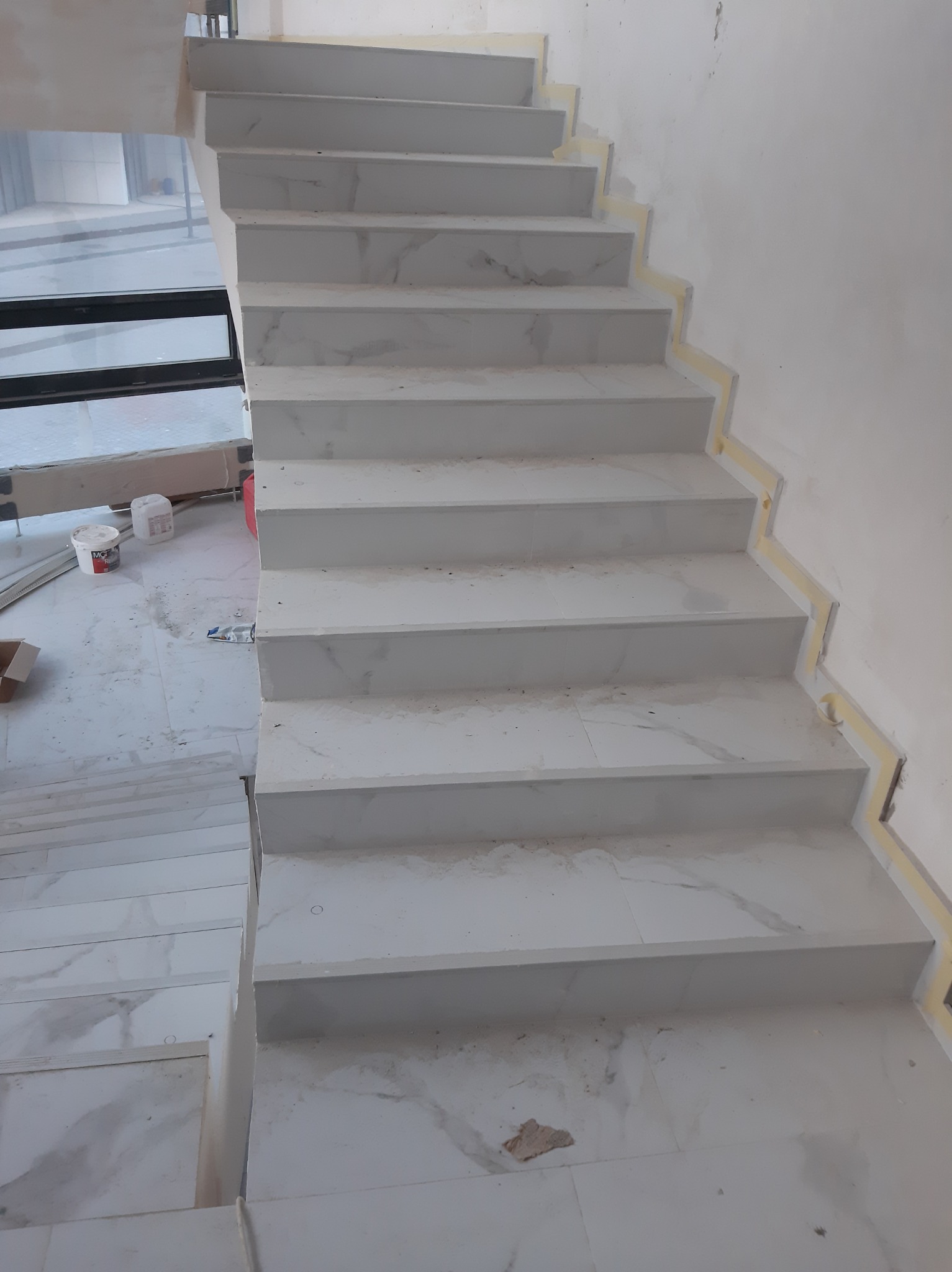 Wyłożone białym marmurem schody wewnętrzne w trakcie wykańczania, zabezpieczone taśmą malarską przy ścianie, widoczne ślady po pracach budowlanych.