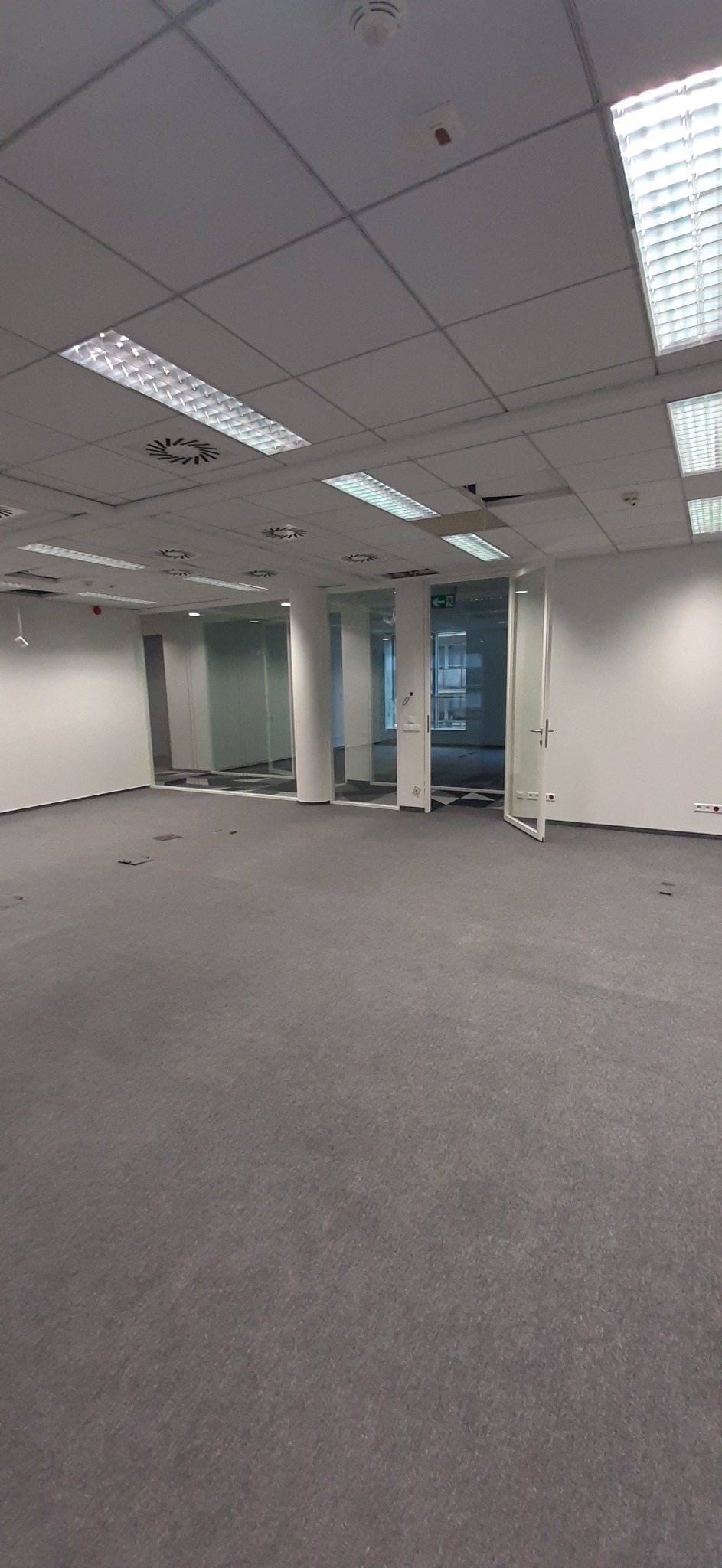 Pusta przestrzeń biurowa z szarą wykładziną, białymi ścianami, podwieszanym sufitem z lampami i systemem wentylacji, oraz szklanymi ścianami działowymi oddzielającymi kolejne pomieszczenia.