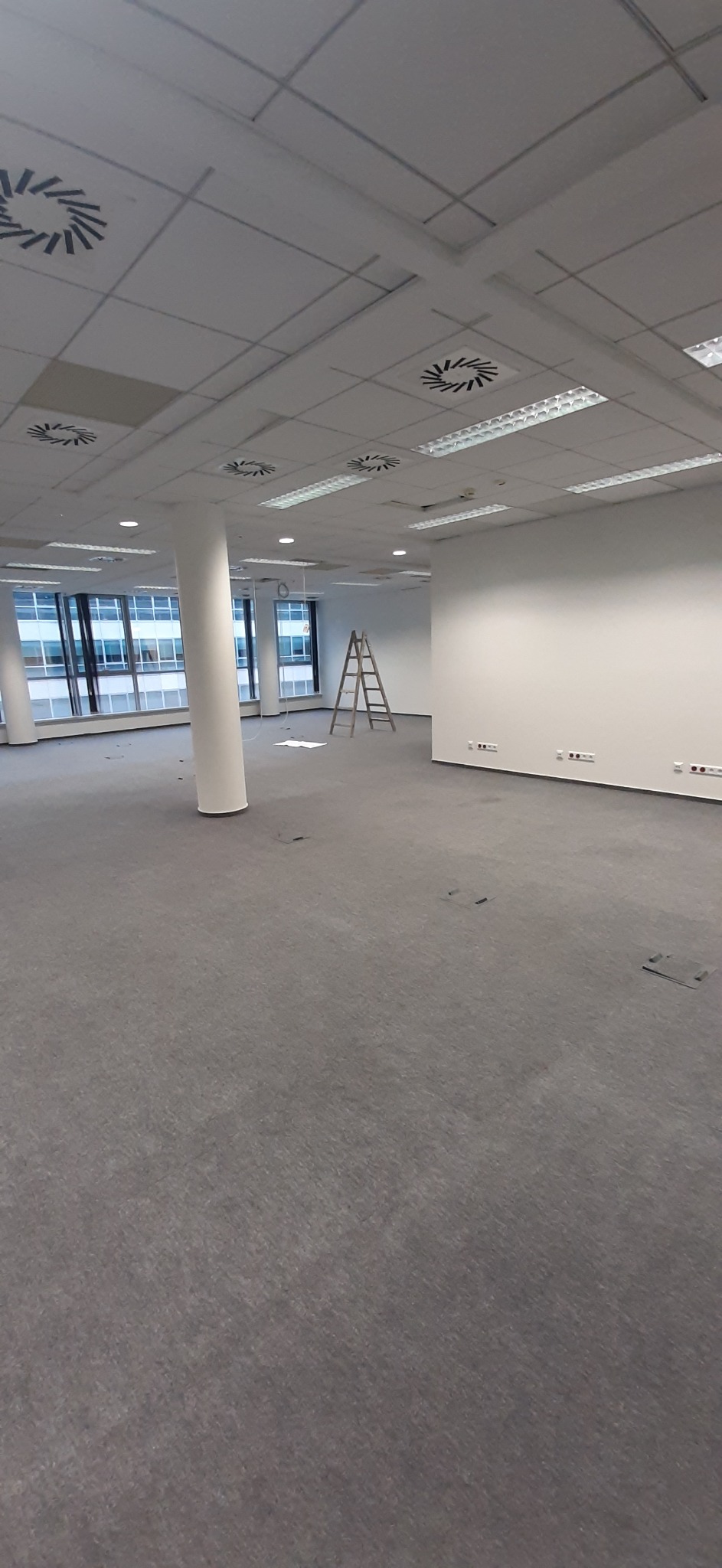 Przestronne, jasne wnętrze biurowe przed wykończeniem, z szarą wykładziną, białą kolumną i aluminiową drabiną, widok na okna z budynkami w tle i białe ściany z gniazdkami elektrycznymi