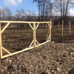 Drewniana brama z siatką na tle zaoranego pola i drzew bez liści, wczesna wiosna.