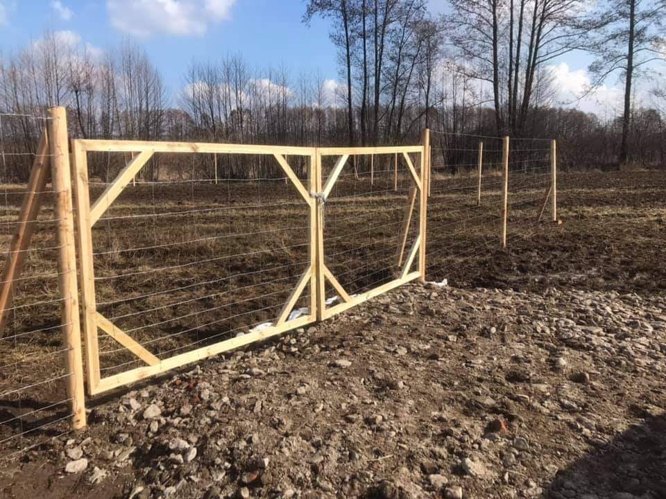 Drewniana brama z siatką w ogrodzeniu polowym, zabezpieczona łańcuchem, z widocznymi słupkami i drutem kolczastym w tle, na nierównym terenie.