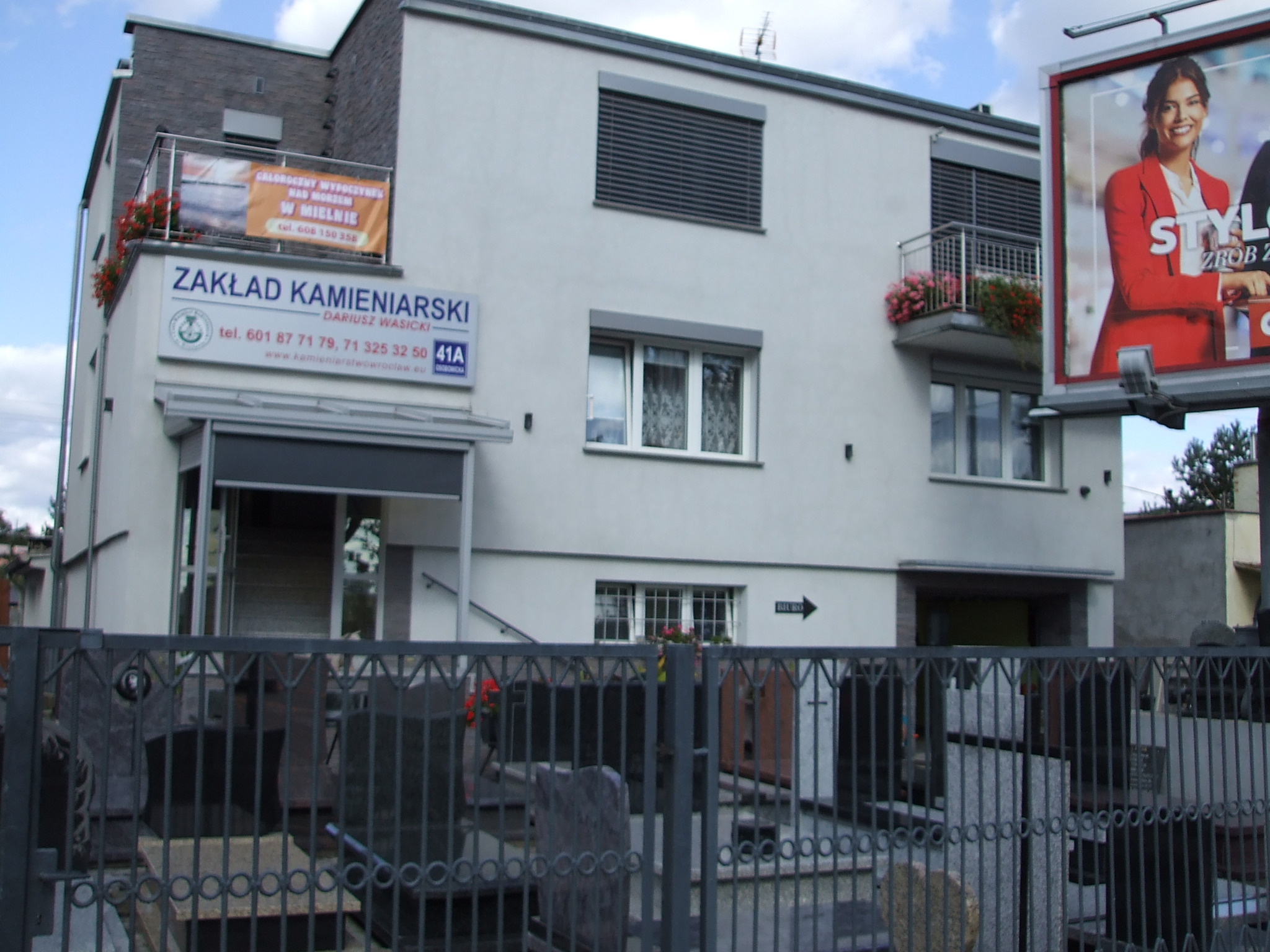 Budynek Zakładu Kamieniarskiego Dariusza Wasickiego z widocznymi nagrobkami za ogrodzeniem, balkonami z kwiatami i reklamą na zewnątrz.