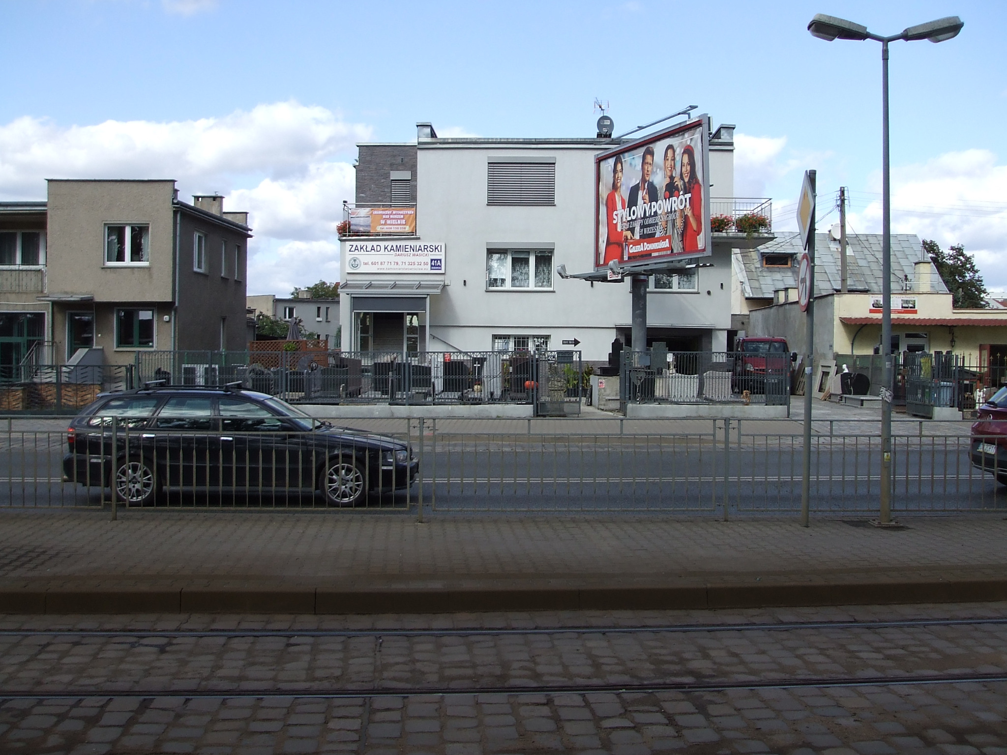 Budynek Zakładu Kamieniarskiego Dariusza Wasickiego z widocznym szyldem i reklamą na balkonie, na pierwszym planie ulica z torami tramwajowymi i zaparkowany czarny samochód.