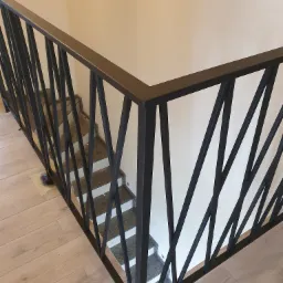 Nowoczesna, czarna balustrada metalowa z geometrycznym wzorem na schodach z szarymi stopniami, widok z góry na jasną podłogę.