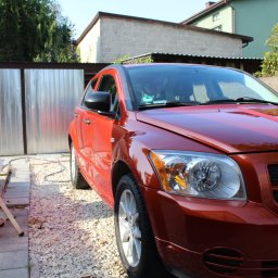 PureSteam - mobilna myjnia parowa - Błyszczący, pomarańczowy samochód Dodge Caliber po umyciu, zaparkowany na kostce brukowej przed garażem z blachy falistej, ze stertą kostki brukowej i aluminiową drabinką w tle.