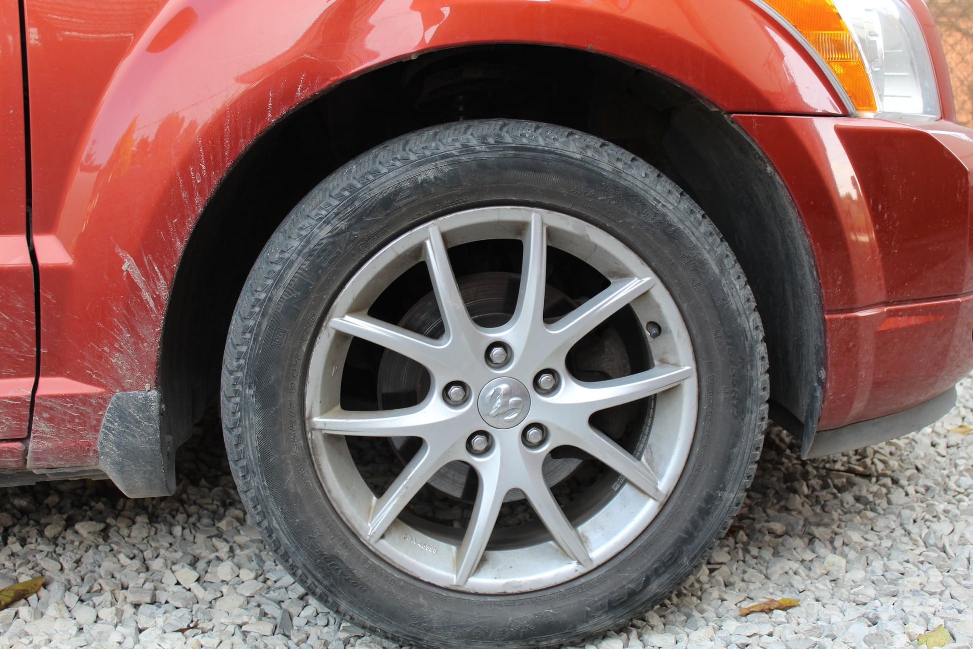 Brudny, pomarańczowy samochód z widocznymi zabrudzeniami na karoserii i oponie, stojący na żwirowym podłożu.