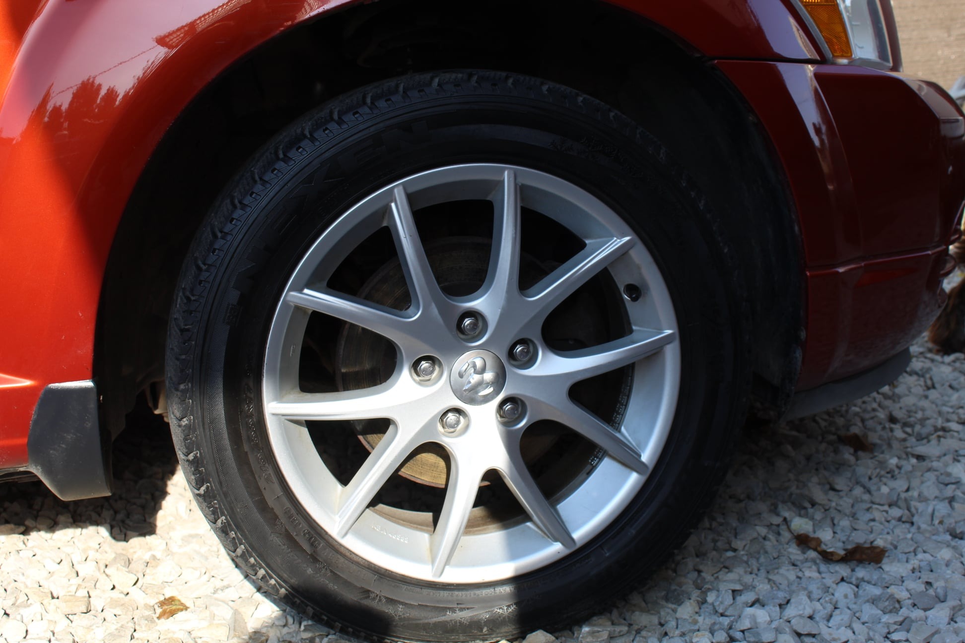 Błyszcząca, aluminiowa felga z oponą na tle czerwonego nadwozia samochodu i żwirowego podłoża, widoczny emblemat producenta na osłonie śruby.