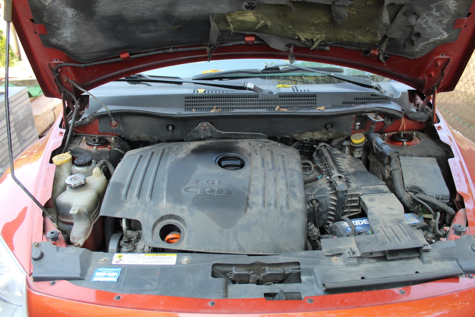 Otwarta maska samochodu z widocznym zakurzonym silnikiem 2.0L CRD i elementami osprzętu, wnętrze maski z widocznymi śladami zużycia.