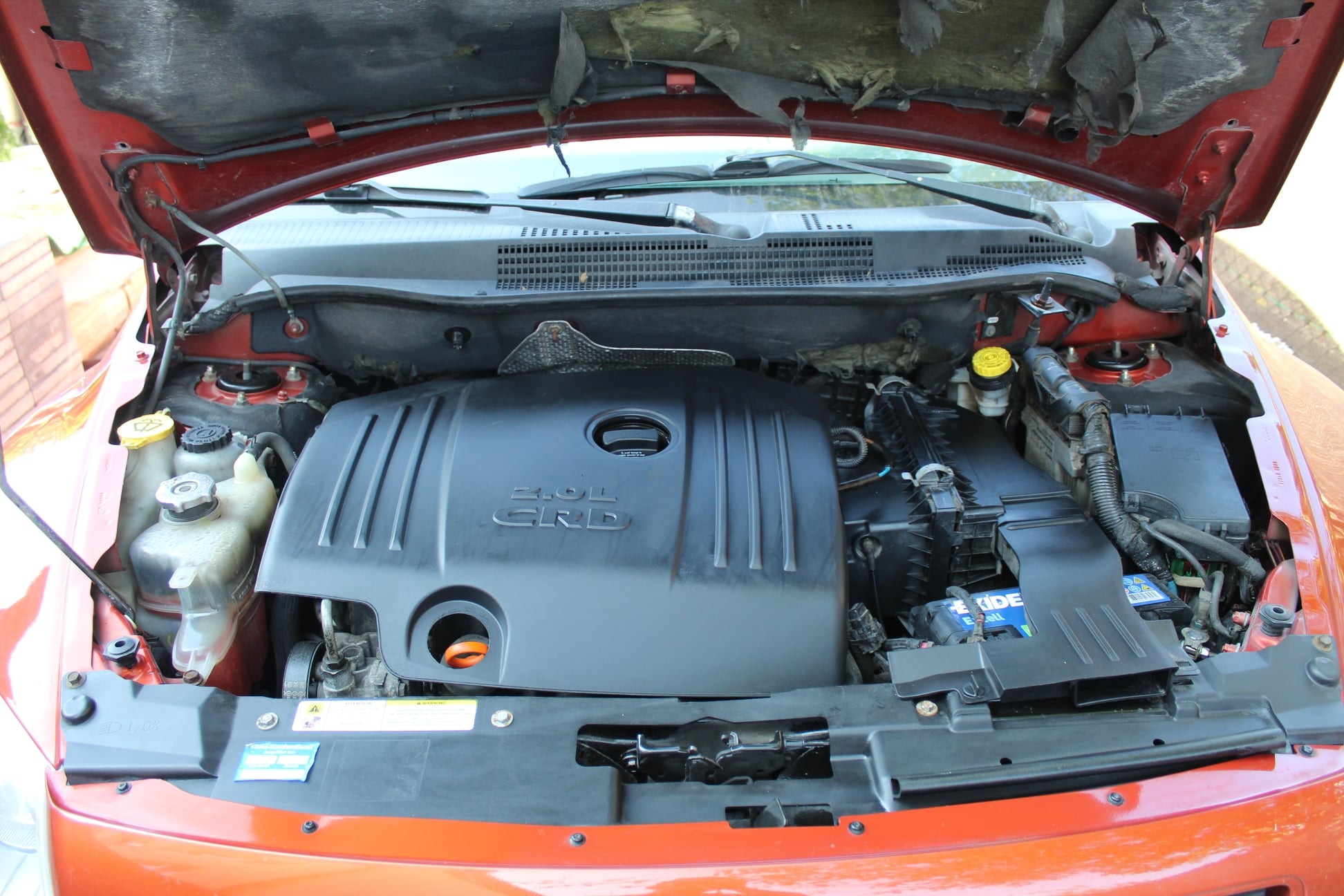Otwarta maska czerwonego samochodu, widoczny silnik 2.0L CRD z elementami plastikowymi i zbiornikami płynów eksploatacyjnych, wnętrze maski częściowo wytarte.