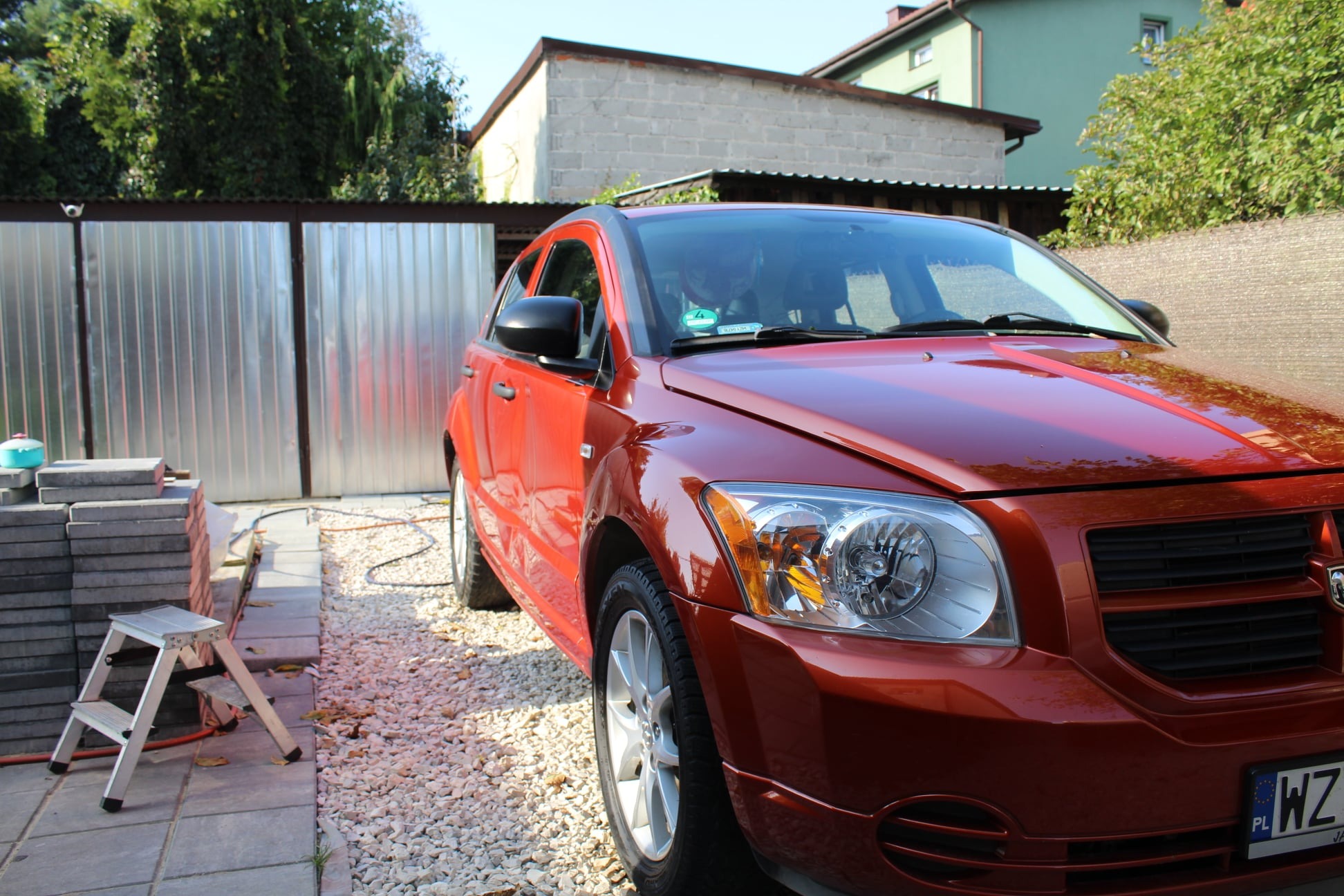Błyszczący, pomarańczowy samochód Dodge Caliber po umyciu, zaparkowany na kostce brukowej przed garażem z blachy falistej, ze stertą kostki brukowej i aluminiową drabinką w tle.