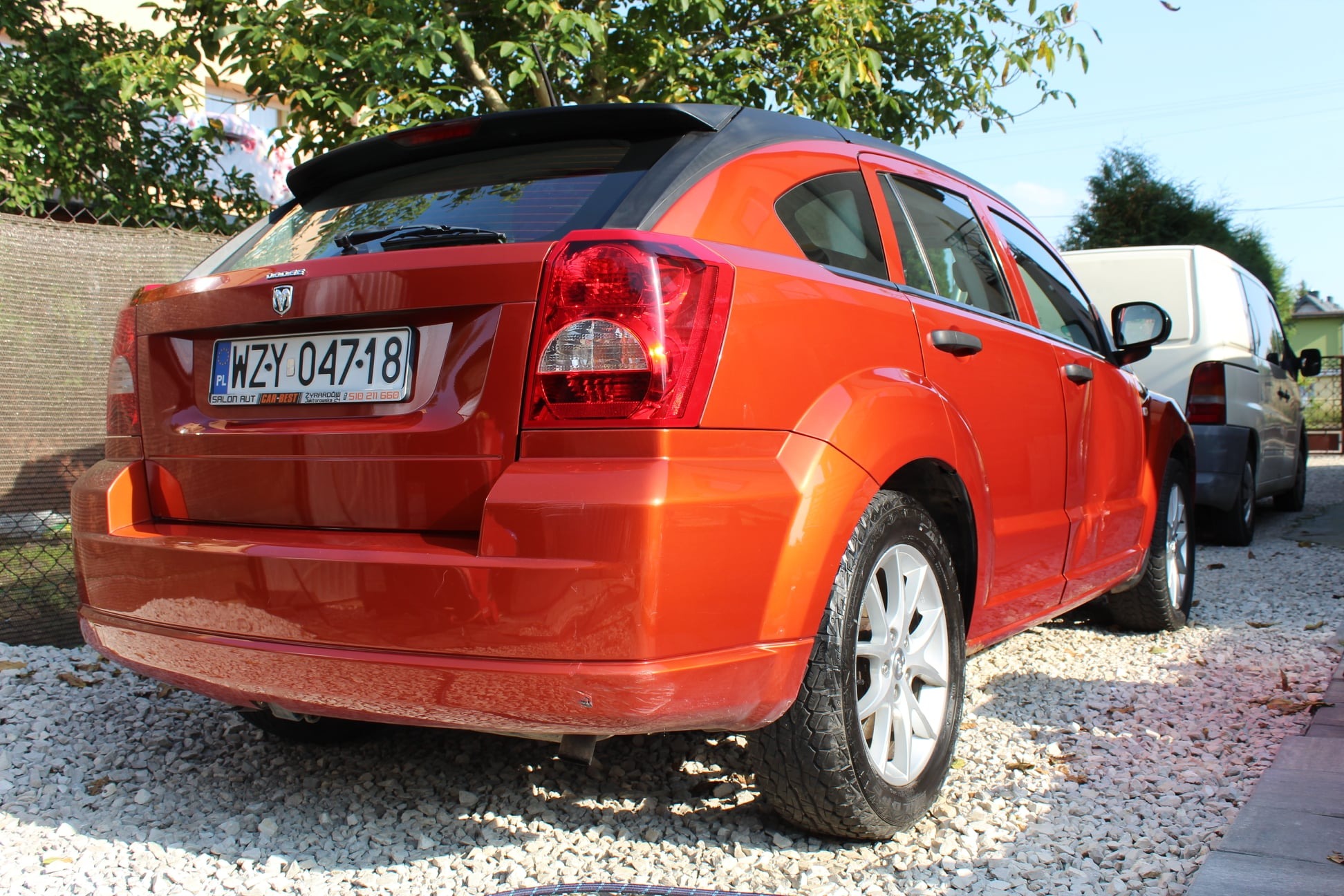 Pomarańczowy samochód Dodge Caliber parkuje na żwirowym podjeździe, widoczny z tyłu, z polską tablicą rejestracyjną i logo dealera samochodowego.