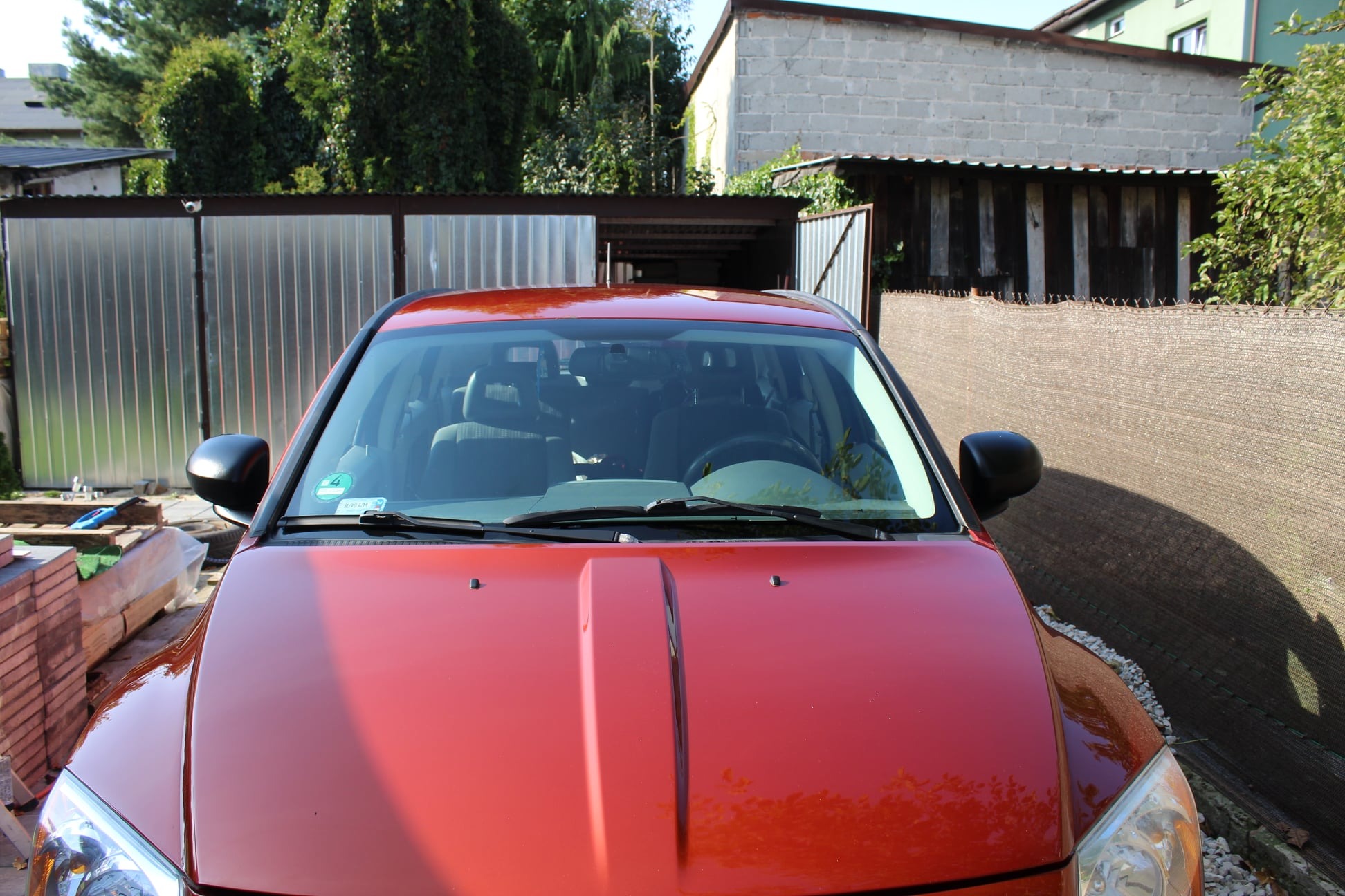 Czerwony samochód zaparkowany na zewnątrz z widoczną stertą cegieł i narzędziami w tle.