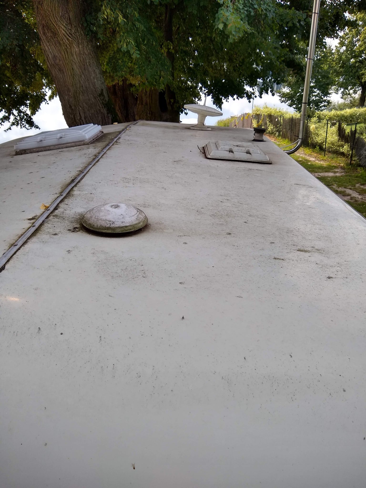 Zabrudzony dach kampera z widocznymi elementami wyposażenia, takim jak okno dachowe, antena i wlot powietrza, na tle drzew i zieleni.