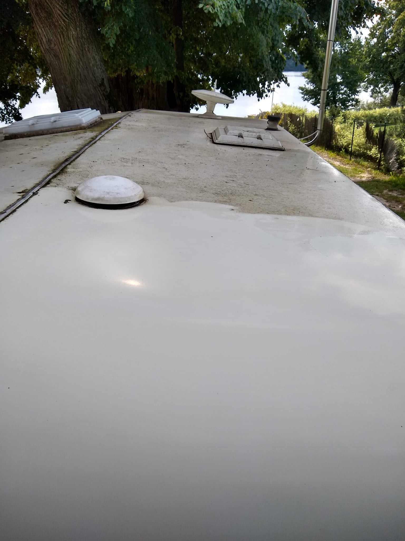 Częściowo umyta, płaska powierzchnia dachu z elementami wyposażenia, widoczne ślady brudu i zacieków, w tle drzewa i jezioro.