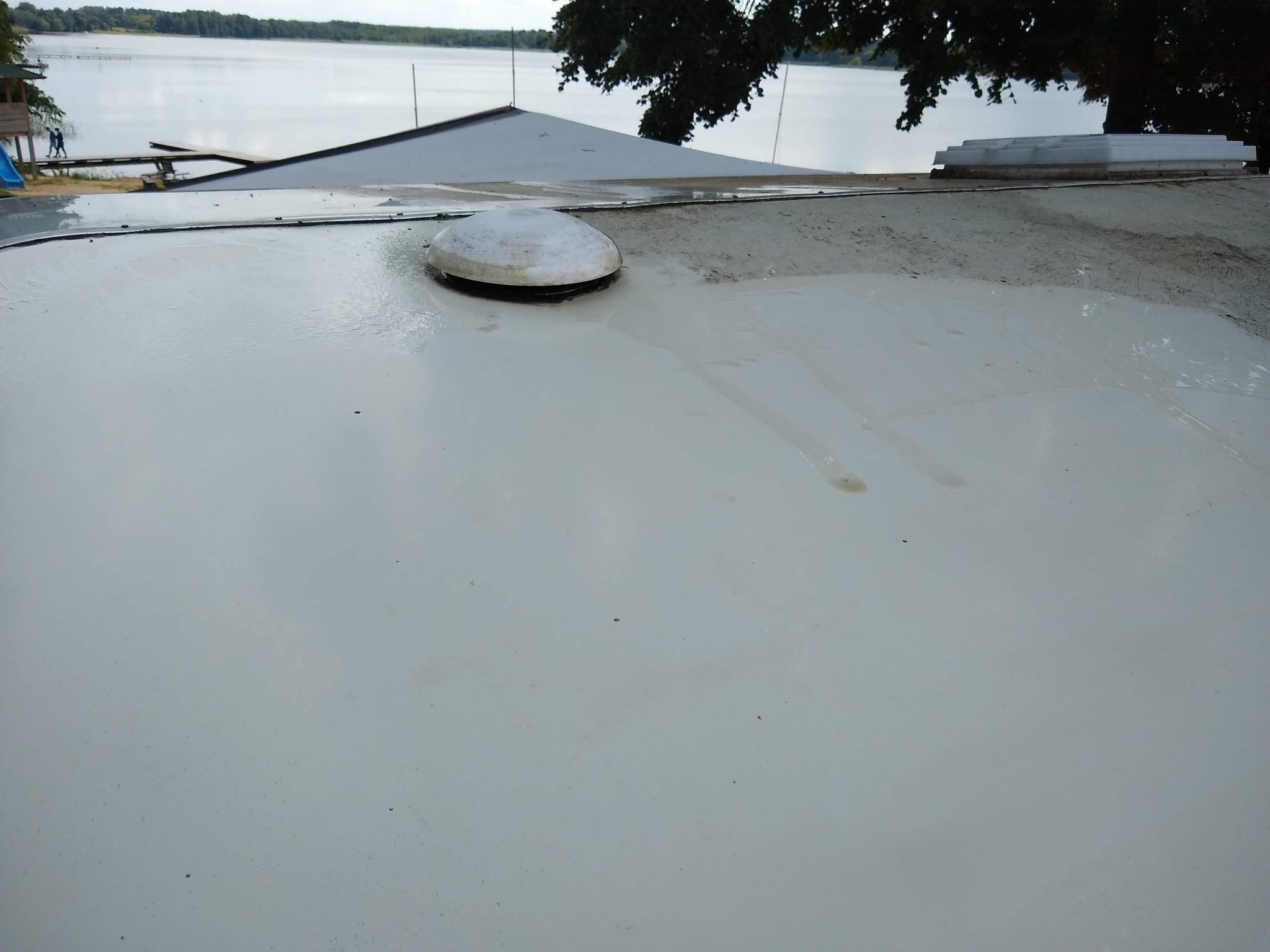 Biała, płaska powierzchnia dachu z widocznym kominkiem wentylacyjnym i śladami po czyszczeniu, w tle jezioro i drzewa.
