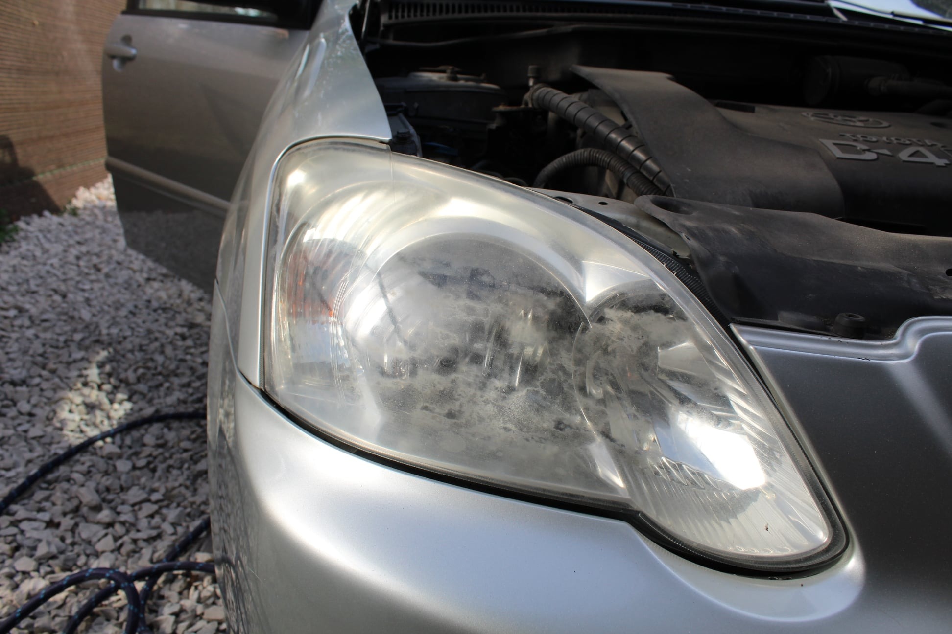 Silnie zabrudzony reflektor srebrnego samochodu z otwartą maską i drzwiami, widoczny na tle żwirowego podłoża.