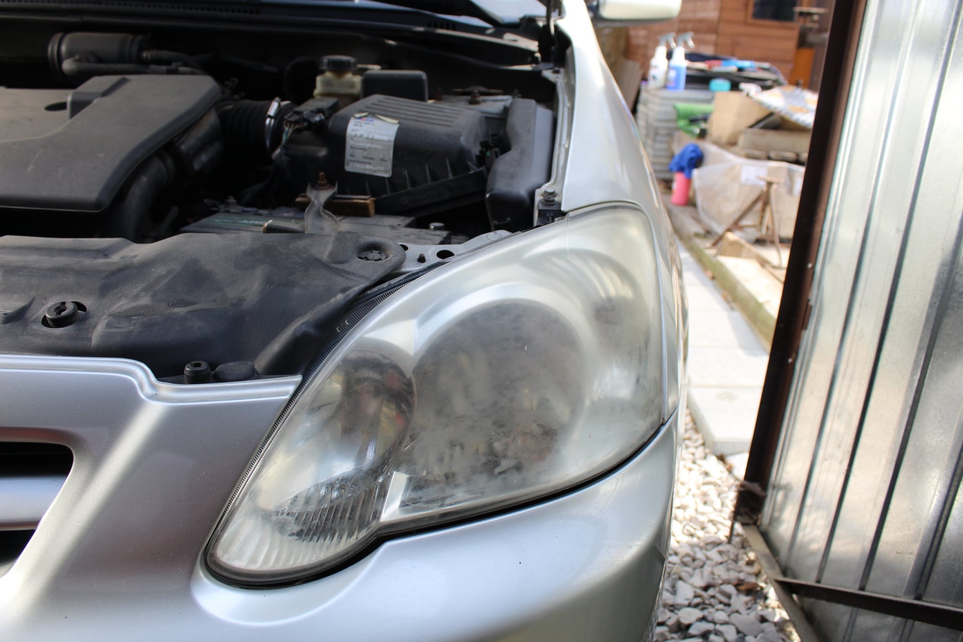 Prawy przedni reflektor srebrnego samochodu z otwartą maską, widoczny silnik w tle, zmatowiały klosz lampy wymagający renowacji.