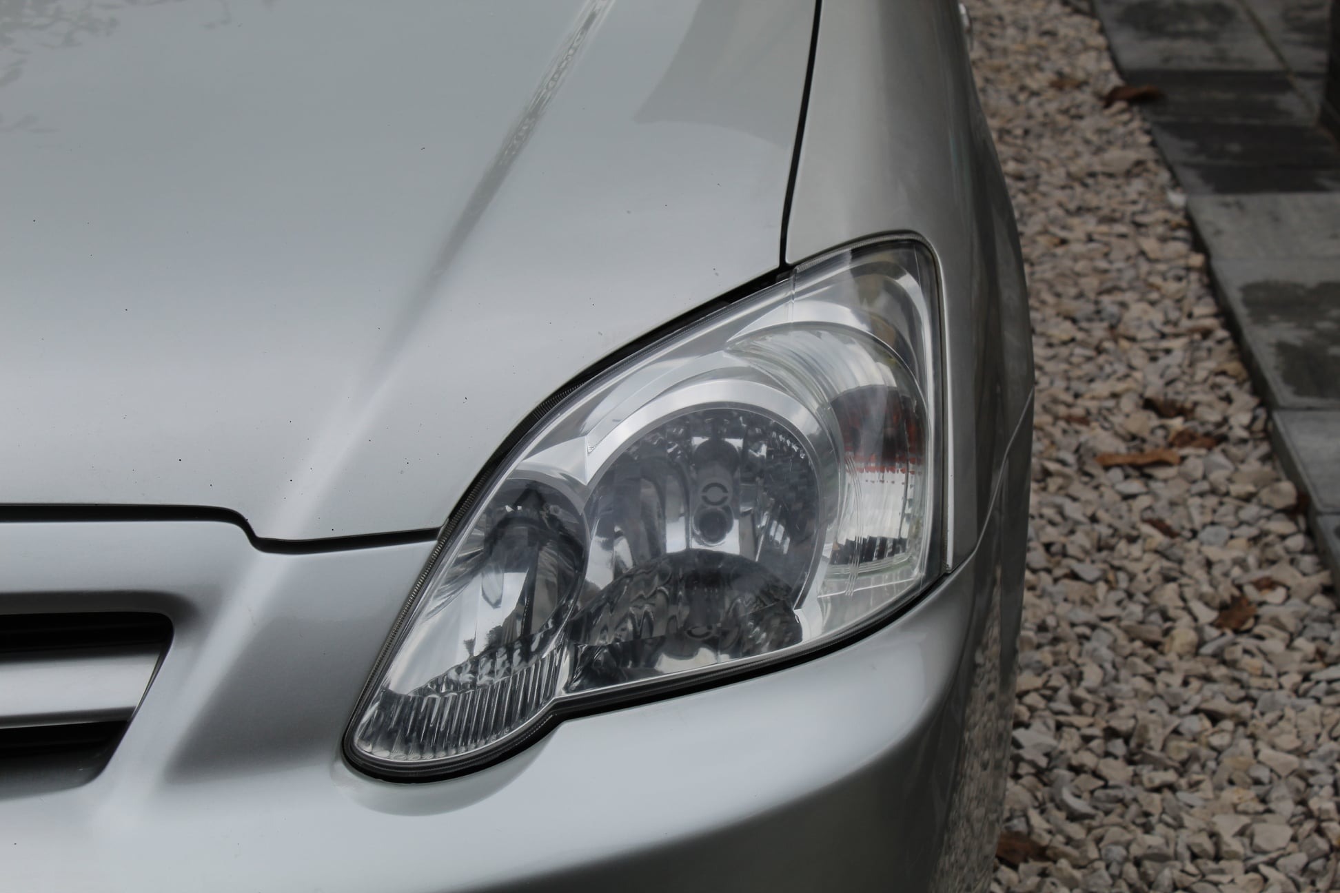 Zbliżenie na reflektor srebrnego samochodu z widocznymi drobnymi zabrudzeniami na karoserii, tło stanowi żwirowa nawierzchnia i fragment betonowego chodnika.