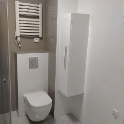 Nowoczesna łazienka z podwieszaną toaletą, grzejnikiem drabinkowym, białą szafką wiszącą i szarymi płytkami podłogowymi, widok z perspektywy kabiny prysznicowej.