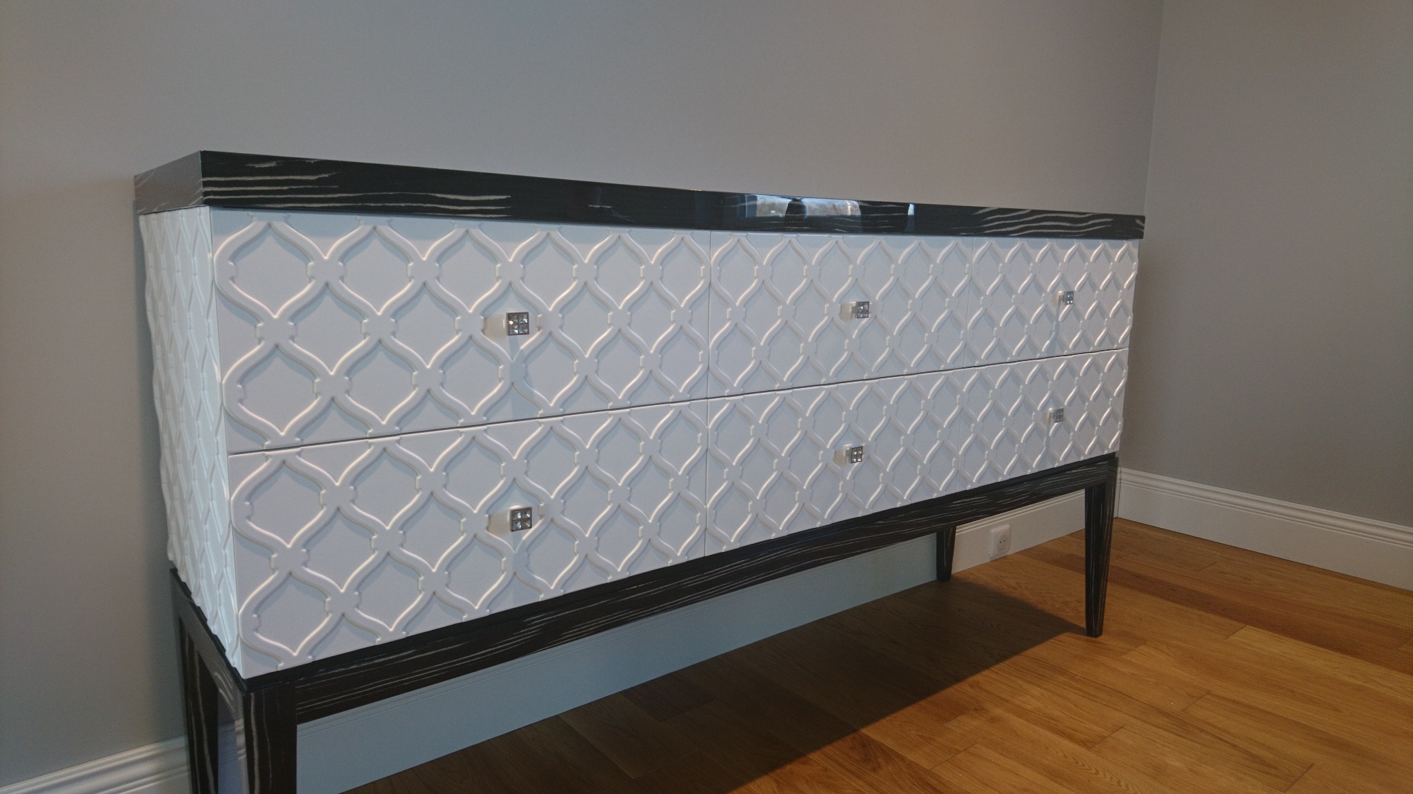 Dwukomorowa komoda z białymi, zdobionymi frontami szuflad i ciemnym, lakierowanym blatem, ustawiona na drewnianej podłodze przy szarej ścianie.