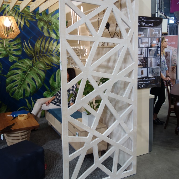 Dekoracyjna ścianka działowa z geometrycznym wzorem, wykonana z jasnego drewna, oddzielająca przestrzeń wypoczynkową z motywem tropikalnym od ekspozycji meblowej.