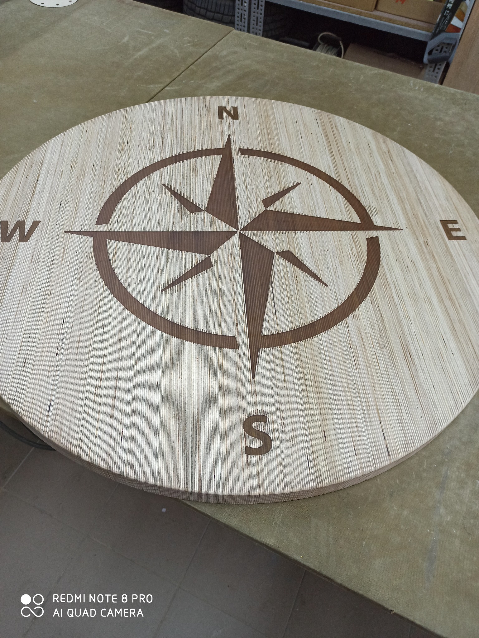 Okrągły blat z jasnego drewna z wygrawerowaną różą wiatrów z oznaczeniami stron świata N, E, S, W.