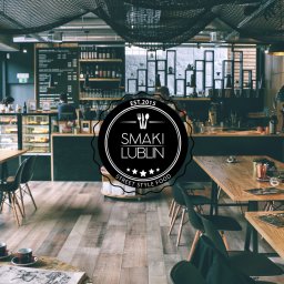 Mistallley - Wnętrze kawiarni Smaki Lublin: drewniane stoły, czarne krzesła, menu na tablicy, ekspozycja ciast, dekoracyjna siatka na suficie i firmowe logo nałożone na obraz