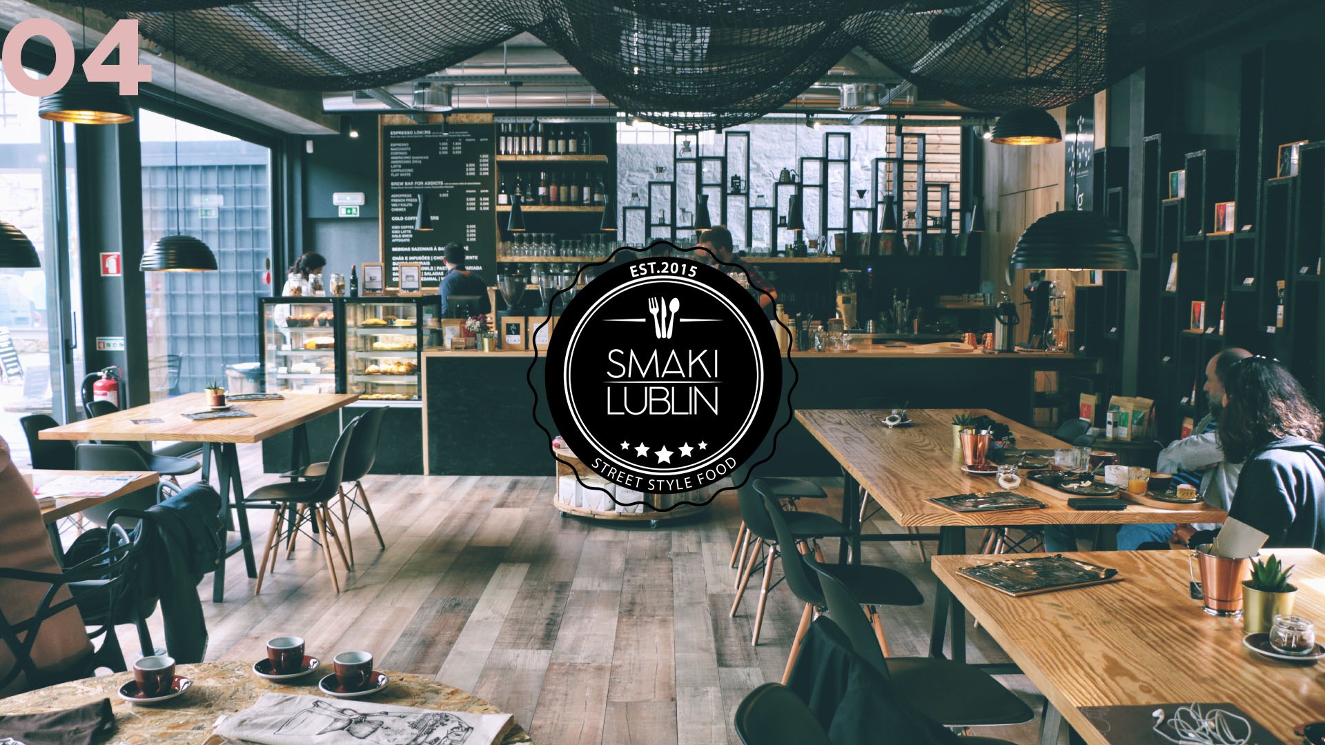 Wnętrze kawiarni Smaki Lublin: drewniane stoły, czarne krzesła, menu na tablicy, ekspozycja ciast, dekoracyjna siatka na suficie i firmowe logo nałożone na obraz