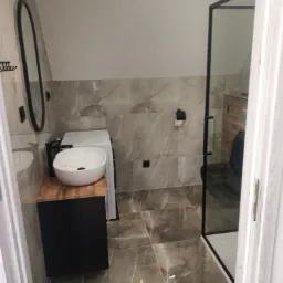 Nowoczesna łazienka z umywalką nablatową na drewnianej szafce, kabiną prysznicową z czarnymi ramami i pralką w tle, wykończona płytkami imitującymi marmur.