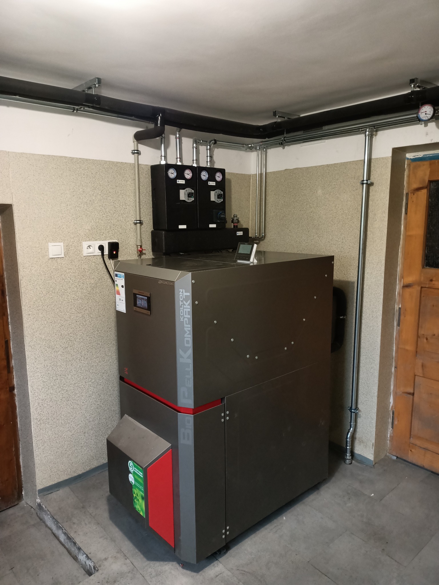 Kompaktowy piec na pellet Kolton w kotłowni, widoczne rury instalacji grzewczej, czujniki i przewody elektryczne.