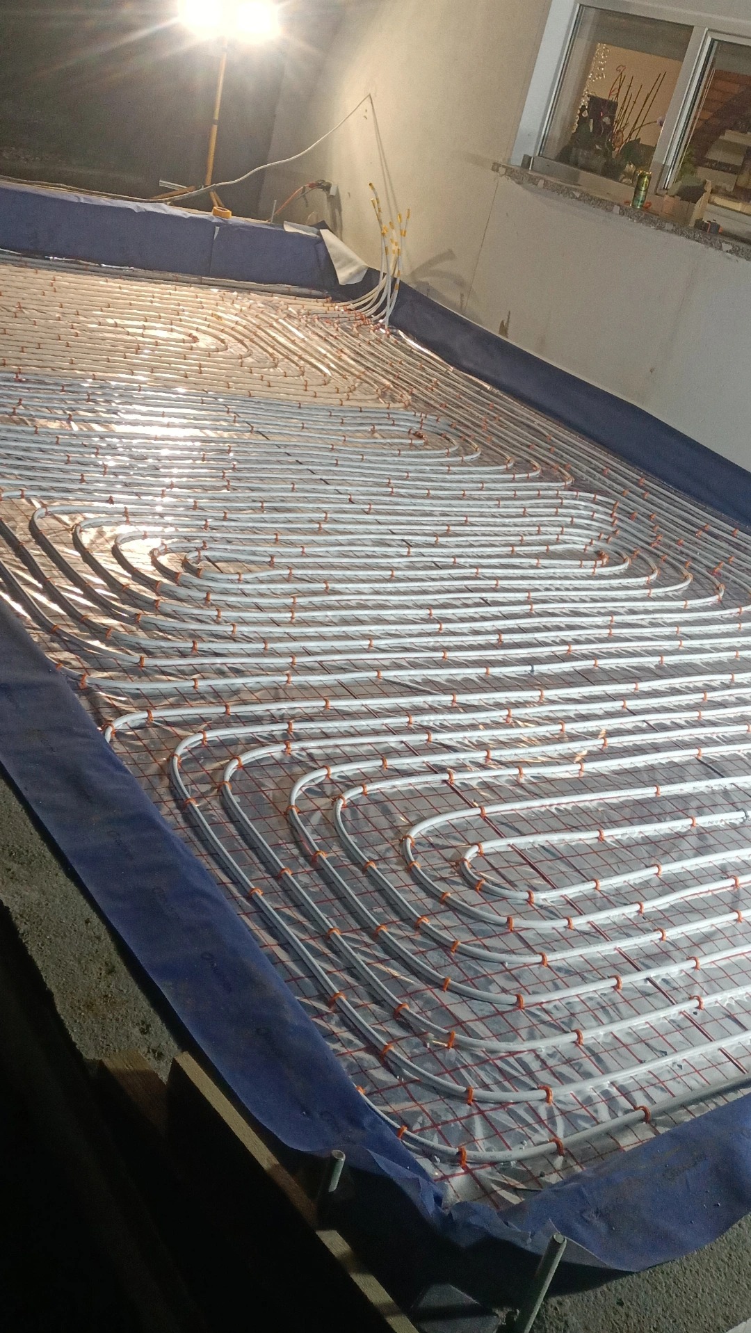 Instalacja ogrzewania podłogowego z rurkami ułożonymi w pętle na folii izolacyjnej, widoczne złącza rur i lampa oświetlająca obszar roboczy.