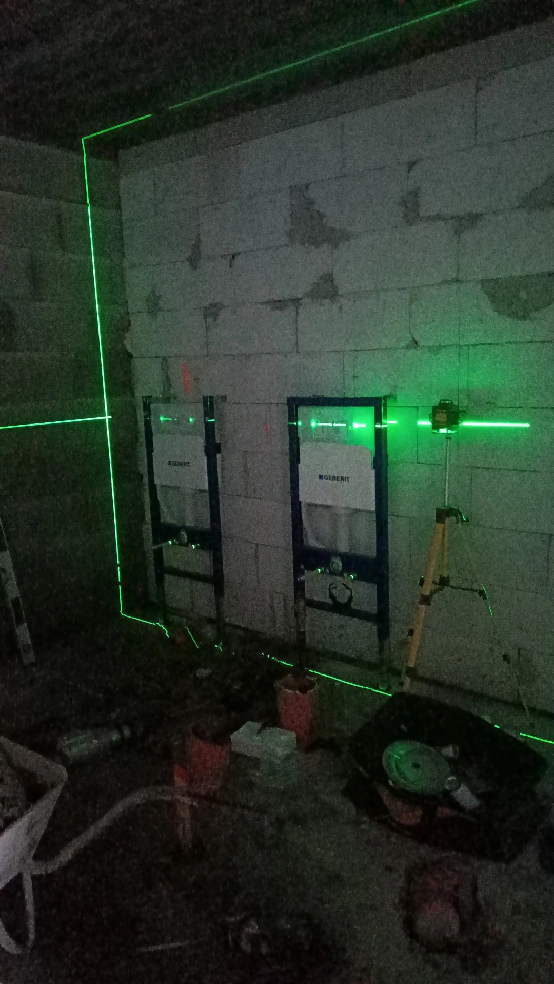 Dwa stelaże podtynkowe Geberit zamontowane na surowej ścianie z betonu komórkowego, poziomowanie za pomocą lasera krzyżowego na statywie, widoczne wiadro i taczka z zaprawą.