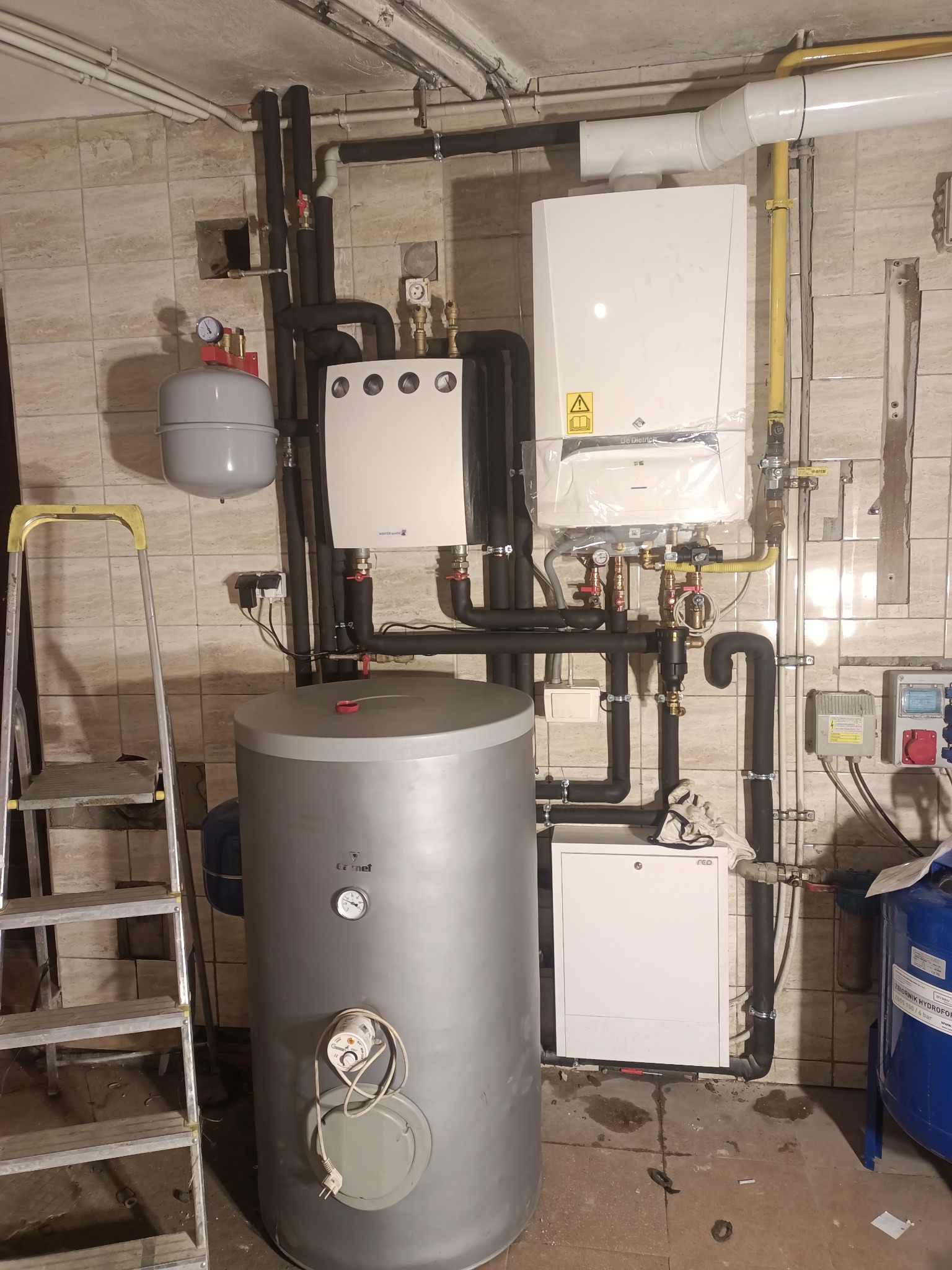 Kotłownia z zamontowanym piecem gazowym, zasobnikiem ciepłej wody i instalacją hydrauliczną, widoczna drabina aluminiowa.