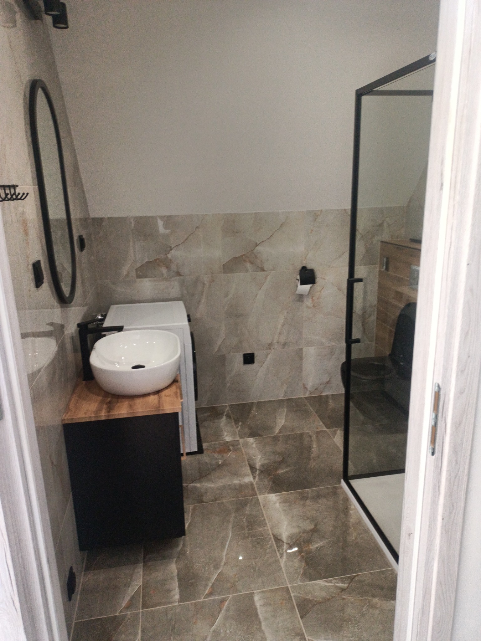 Nowoczesna łazienka z umywalką nablatową na drewnianej szafce, kabiną prysznicową z czarnymi ramami i pralką w tle, wykończona płytkami imitującymi marmur.