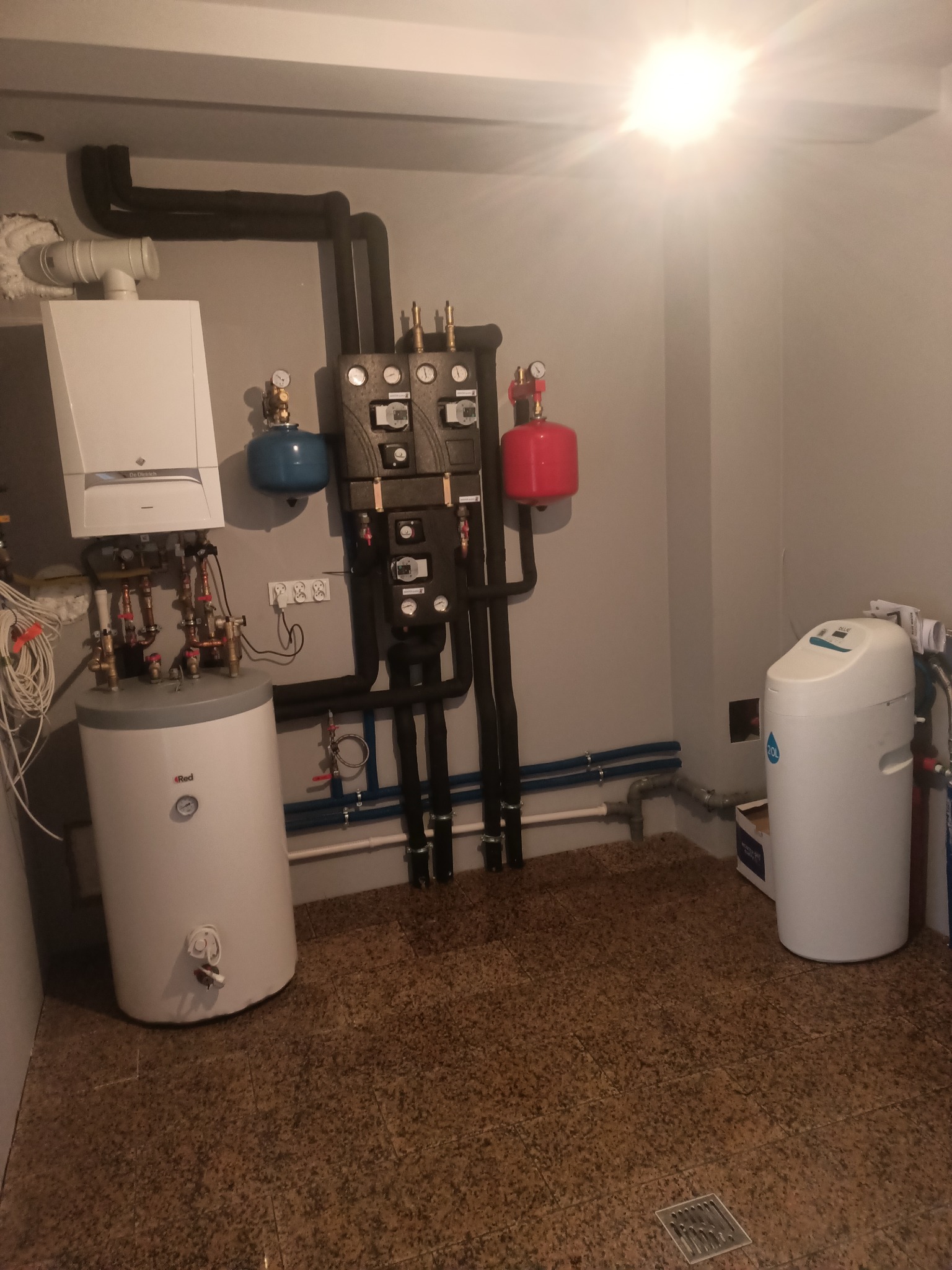 Kompaktowa kotłownia z białym piecem gazowym, zasobnikiem CWU, rozdzielaczami, czarnymi rurami izolowanymi termicznie oraz filtrem wody na brązowej posadzce z kratką ściekową.