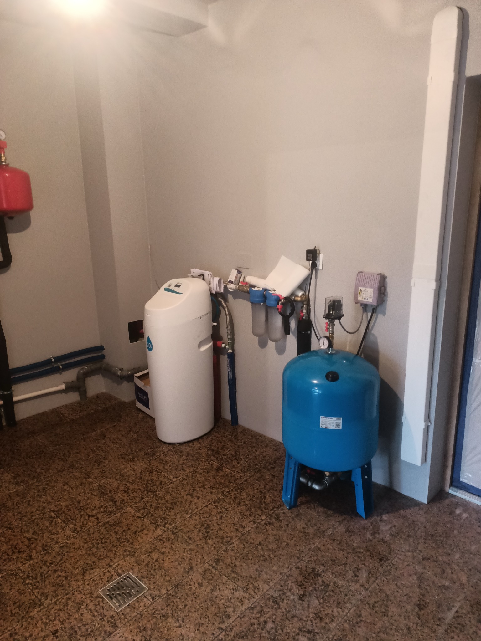 Zestaw do uzdatniania wody z niebieskim zbiornikiem hydroforowym Aquasystem, filtry i biały zmiękczacz wody w pomieszczeniu technicznym z brązową podłogą z terakoty.