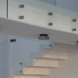 Nowoczesne schody z białymi stopniami i szklaną balustradą mocowaną na metalowe śruby, oświetlenie punktowe pod sufitem.