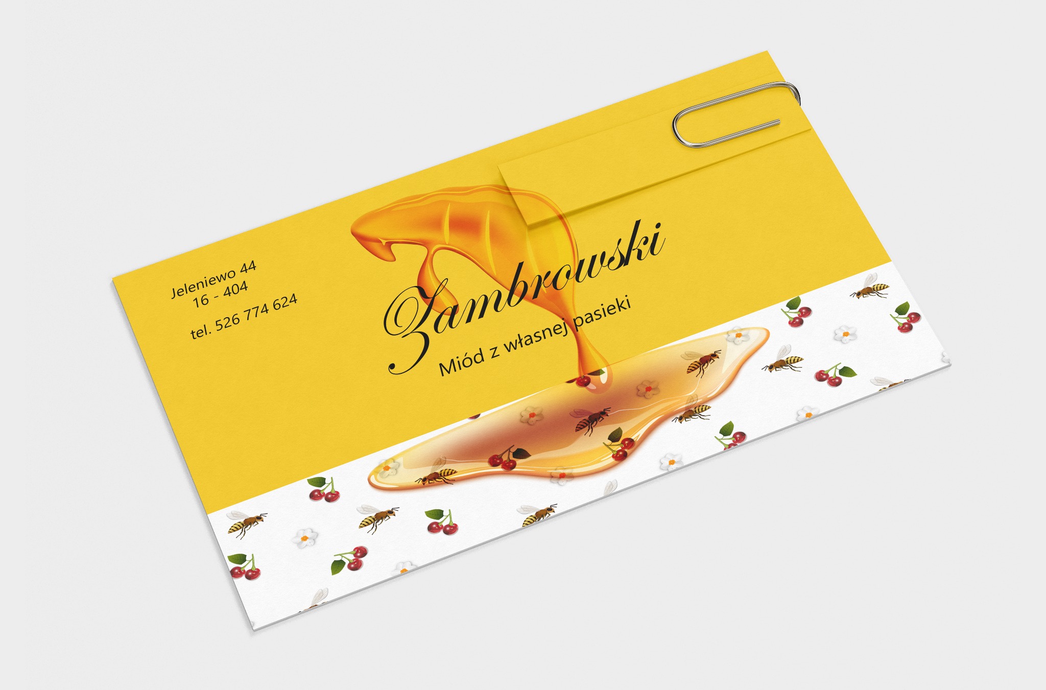 Żółta wizytówka z motywem miodu, wisien i pszczół, spięta srebrnym spinaczem biurowym. Wizytówka prezentuje nazwę firmy 'Zambrowski' oraz dopisek 'Miód z własnej pasieki', a także dane kontaktowe.
